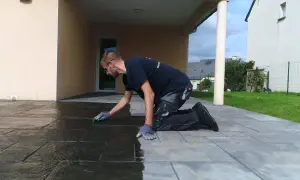 Un homme agenouillé effectue le nettoyage de pierre naturelle extérieur avec une éponge, redonnant son éclat à la terrasse.