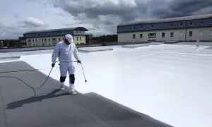 Un ouvrier applique une peinture réflective toiture blanche sur un toit plat. Le ciel est couvert de nuages sombres.