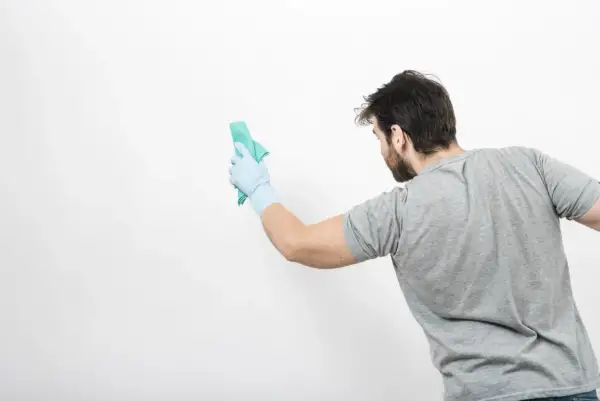 Un homme en gant bleu nettoie un mur blanc avec un chiffon vert. Il montre comment nettoyer un mur blanc sans enlever la peinture.