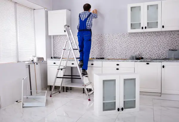 Un homme en salopette bleue est sur un escabeau, prêt à fixer un meuble au mur. La cuisine est en cours d'installation.