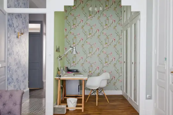 Un pan de mur de salon est habillé d'un papier peint floral aux fleurs roses sur fond vert pâle. Un bureau et une chaise blanche complètent l'espace.